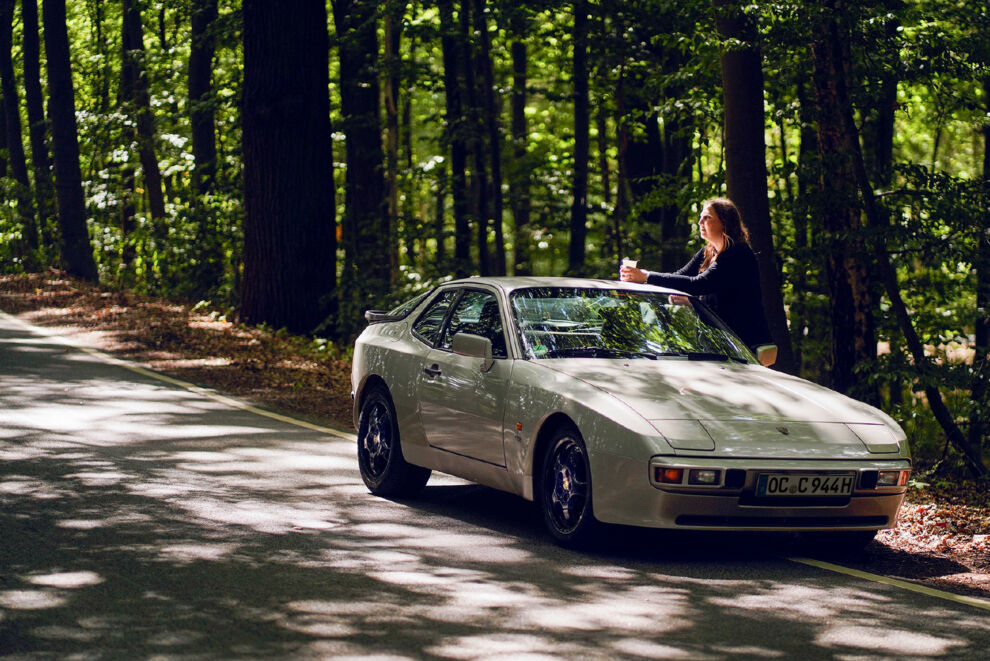 Frau steht neben einem grauen Porsche in einer Waldumgebung